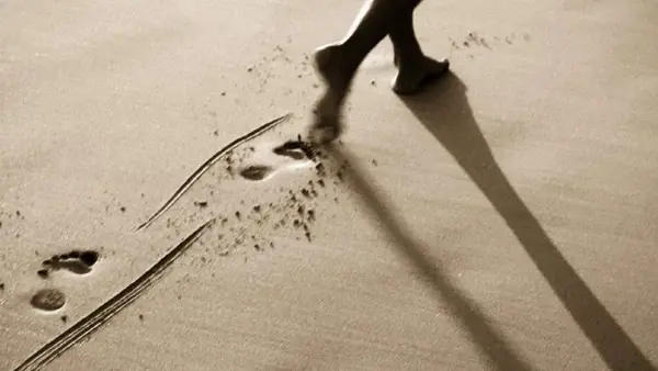 Pegadas na areia e a sombra de uma figura caminhando, em tons sépia